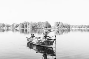 A small fishing boat floats on a calm lake with trees reflecting in the monochrome water.