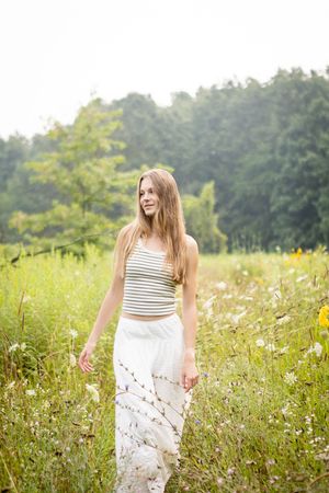 A woman in a striped crop top and white skirt walks through a sunny wildflower meadow on a summer afternoon.