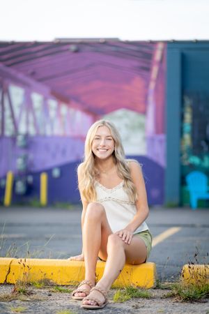 Casual summer portrait of person sitting on yellow curb near colorful wall.