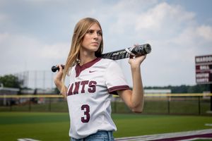 A softball player in a white jersey with maroon accents stands ready at bat on a sunny day at the field.