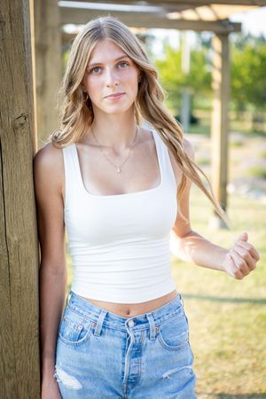 Woman in white crop top and ripped blue jeans poses against wooden posts in outdoor summer setting.