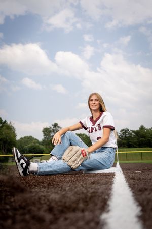 A baseball player sits casually on a dirt field baseline wearing a jersey, jeans and sneakers on a sunny day.