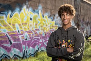 Smiling person in a hoodie standing in front of colorful graffiti wall with crossed arms and curly hair.