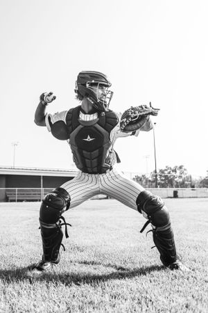 Baseball catcher in protective gear kneeling in ready position on baseball field in black and white photograph.