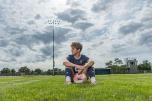 A football player sits cross-legged on a grassy field with stadium lights and cloudy skies in the background.