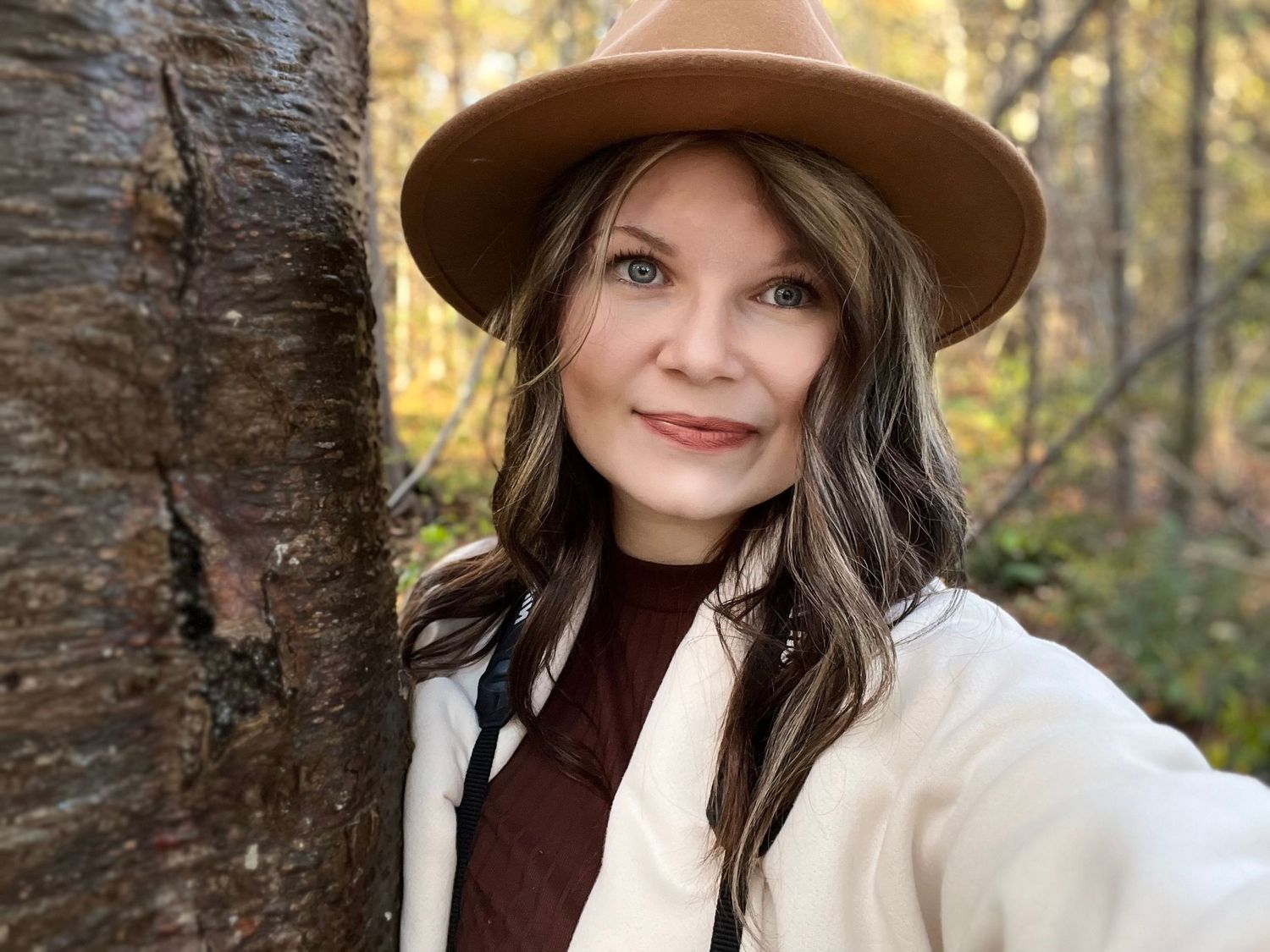 Woman in tan hat and white coat leans against tree trunk in autumn forest selfie portrait.