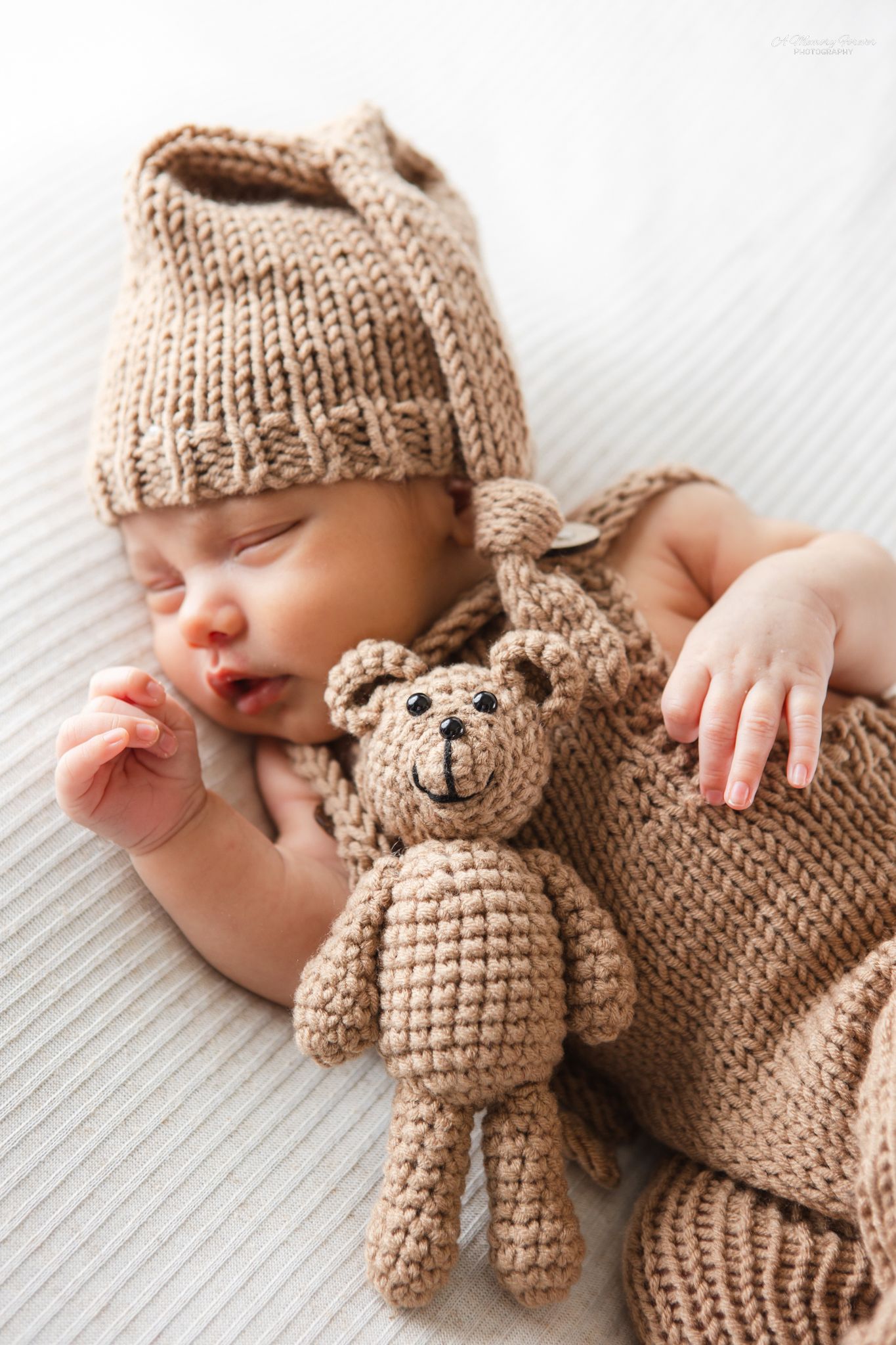 Sleeping newborn baby wearing a knit beige hat and outfit, cuddling a small crocheted teddy bear on a white blanket.