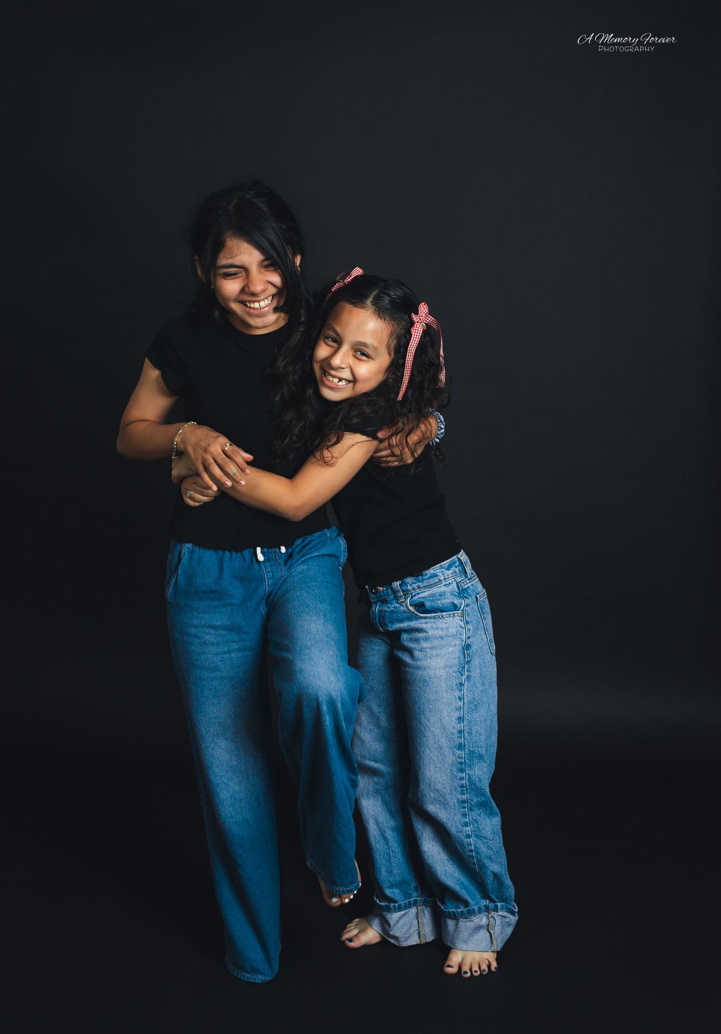 A woman and young girl in matching black tops and jeans hugging and laughing against a dark background.