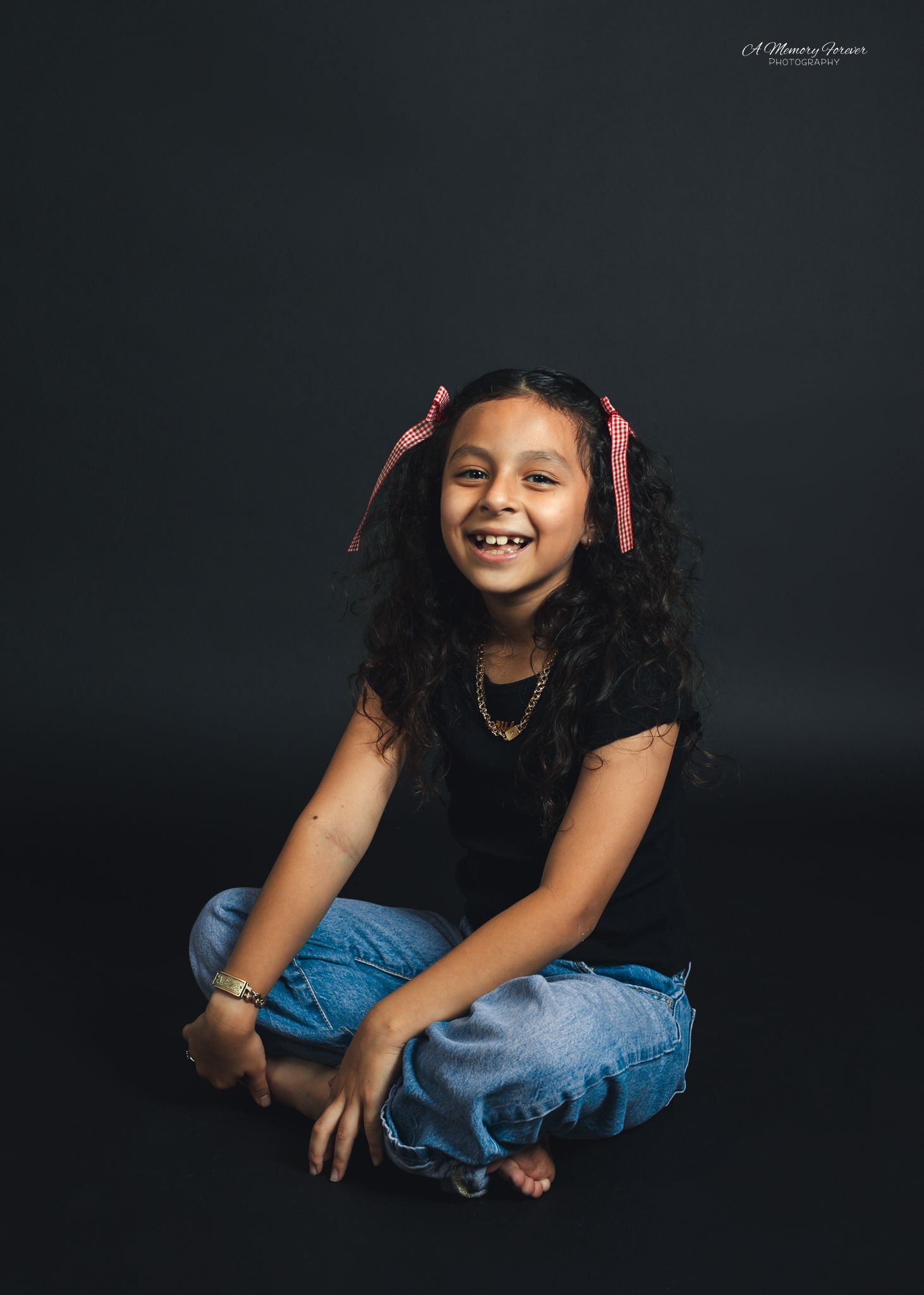 Young girl with curly hair and pink ribbons smiles brightly, sitting cross-legged in jeans and black top against dark backdrop.