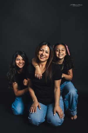 A mother and two daughters smile together in matching black tops and jeans against a dark studio background.