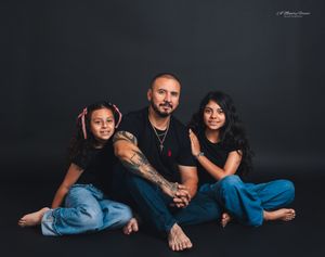 A man with tattoos sits on the floor with two young girls, all wearing black tops, posing for a family portrait.