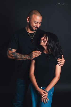 A couple in matching black outfits share a tender moment, the man with tattoos and a cross necklace embracing a smiling woman.