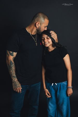 Tattooed man in black t-shirt kissing young girl's head, both smiling, studio portrait with dark background.