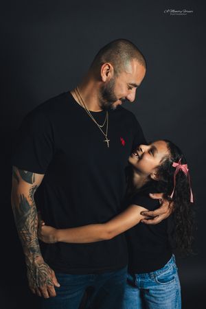 A tattooed father in a black t-shirt and gold cross necklace shares a tender moment with his young daughter in a studio portrait.