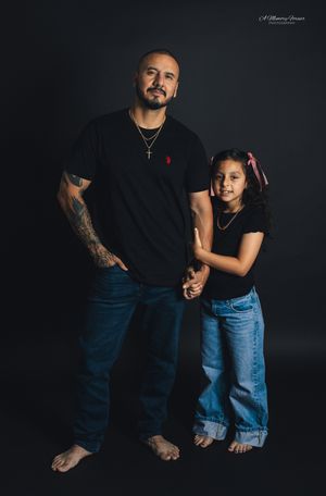 A man and young girl in matching black shirts and jeans pose together barefoot against a dark studio background.