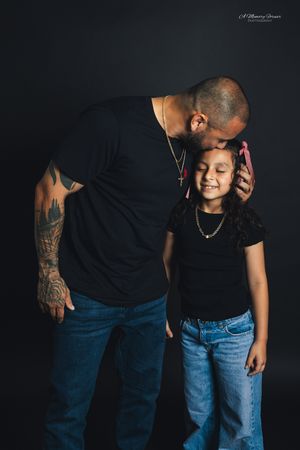 A tattooed father kisses his young daughter on the head as she smiles, both wearing black shirts and jeans.