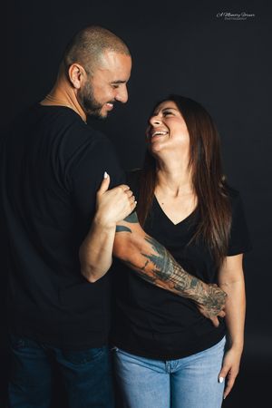 Couple in black t-shirts laughing together, woman with tattoos embracing man against dark studio background.