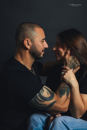A tattooed couple in black shirts share an intimate moment, gazing into each other's eyes against a dark background.