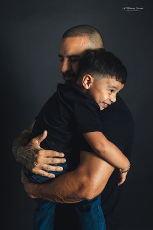 A tattooed father tenderly holds his smiling young son in a warm embrace against a dark studio background.