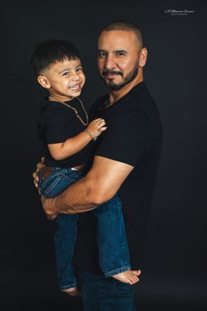 A bald man with a beard holds a smiling young boy, both wearing black shirts and jeans against a dark background.