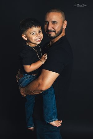 A bald man with a beard holds a smiling young boy, both wearing black shirts and jeans against a dark background.