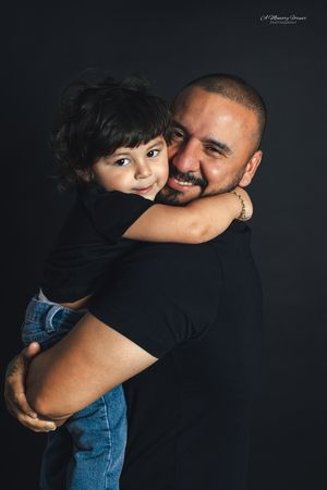 A smiling man with a shaved head holds a young child in a warm embrace against a dark studio background.