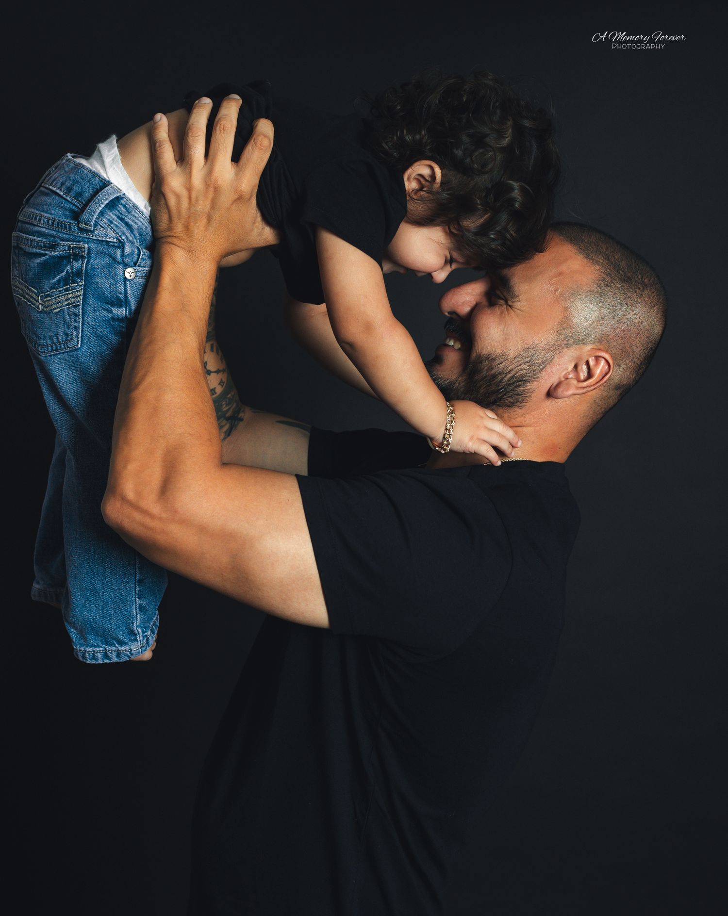 A man in a black shirt lifts a young child close for a tender forehead touch against a dark background.