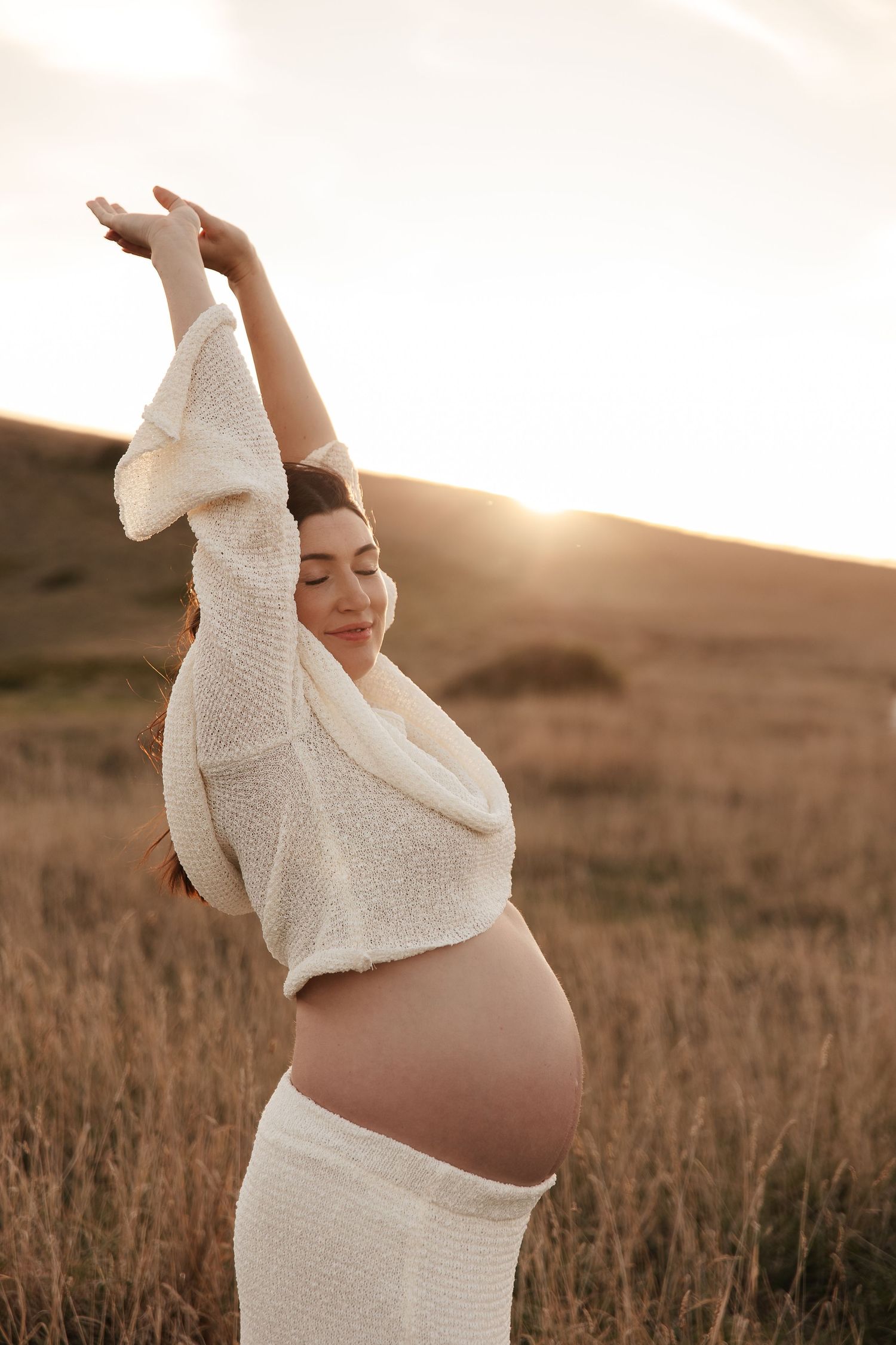 Expectant mother in pink dress poses on beach at golden hour.