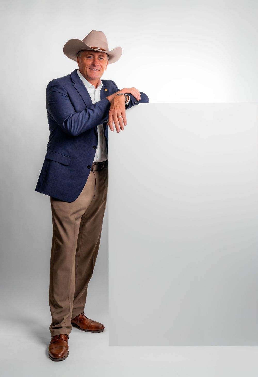 A person in a navy blazer and cowboy hat leans casually against a white wall while wearing brown dress shoes and khakis.