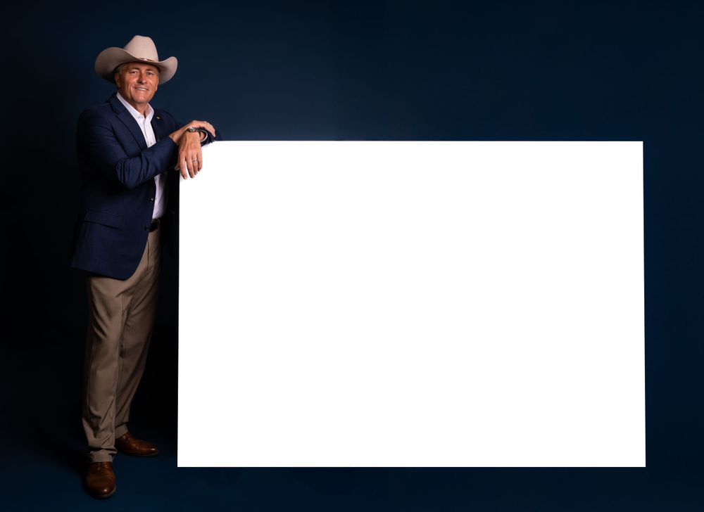 A cowboy in a blue blazer stands beside a large white blank sign against a dark background.
