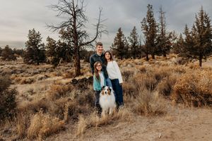 A couple and their dog pose together in a desert landscape with sage brush and evergreen trees at sunset.