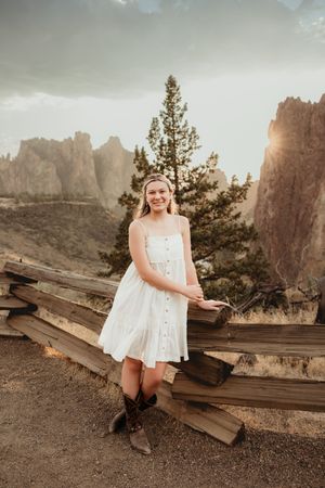 A person in a white dress and cowboy boots sits on wooden railroad ties with scenic mountain cliffs in the background.