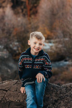 Child in a patterned winter sweater sits on rocky terrain during autumn with warm lighting and blurred background.