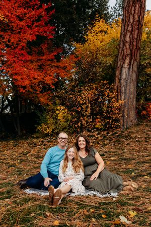 A family sits together on fallen pine needles surrounded by vibrant red and yellow autumn foliage in a forest setting.