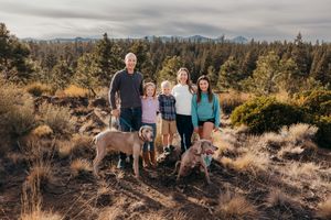 A family and their dogs pose together on a scenic hiking trail with mountains and forest in the background.