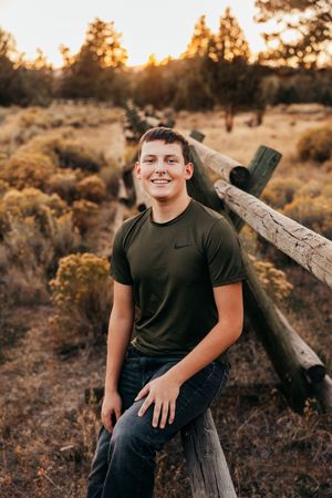 A young person in a black t-shirt poses on wooden logs at sunset in a desert landscape.