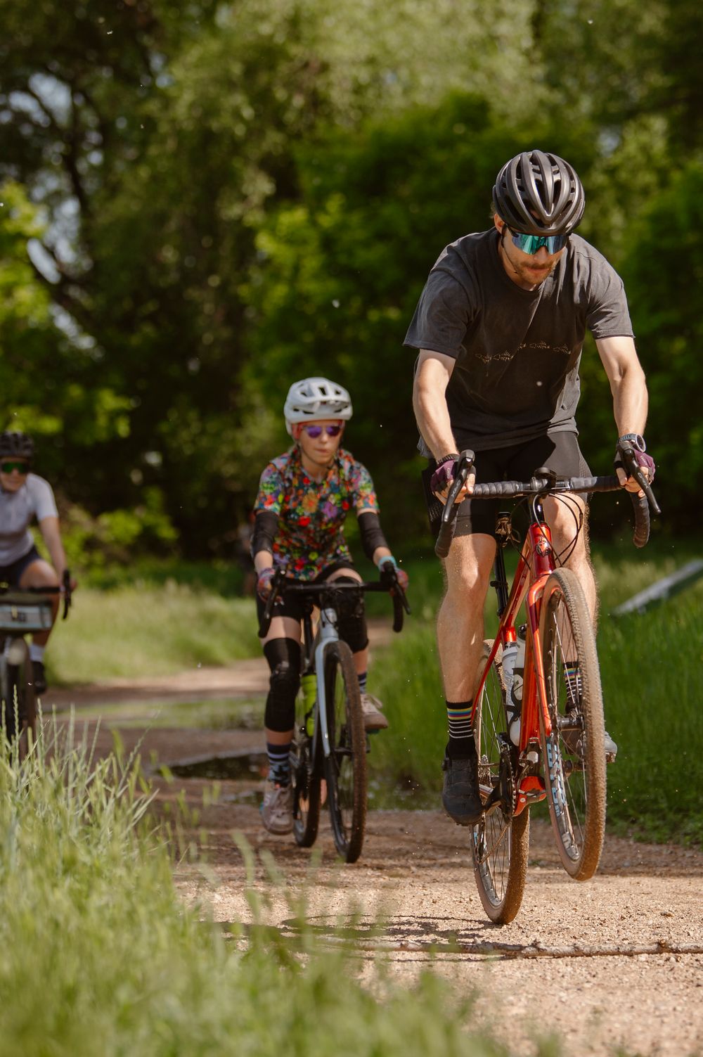 Mountain bikers ride in sequence on a dirt trail through green foliage on a sunny day.