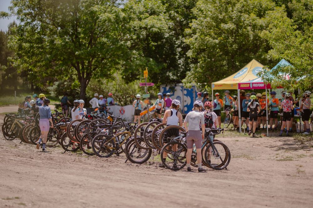 A group of mountain bikes parked at a dirt lot gathering spot with tents and people during an outdoor event.