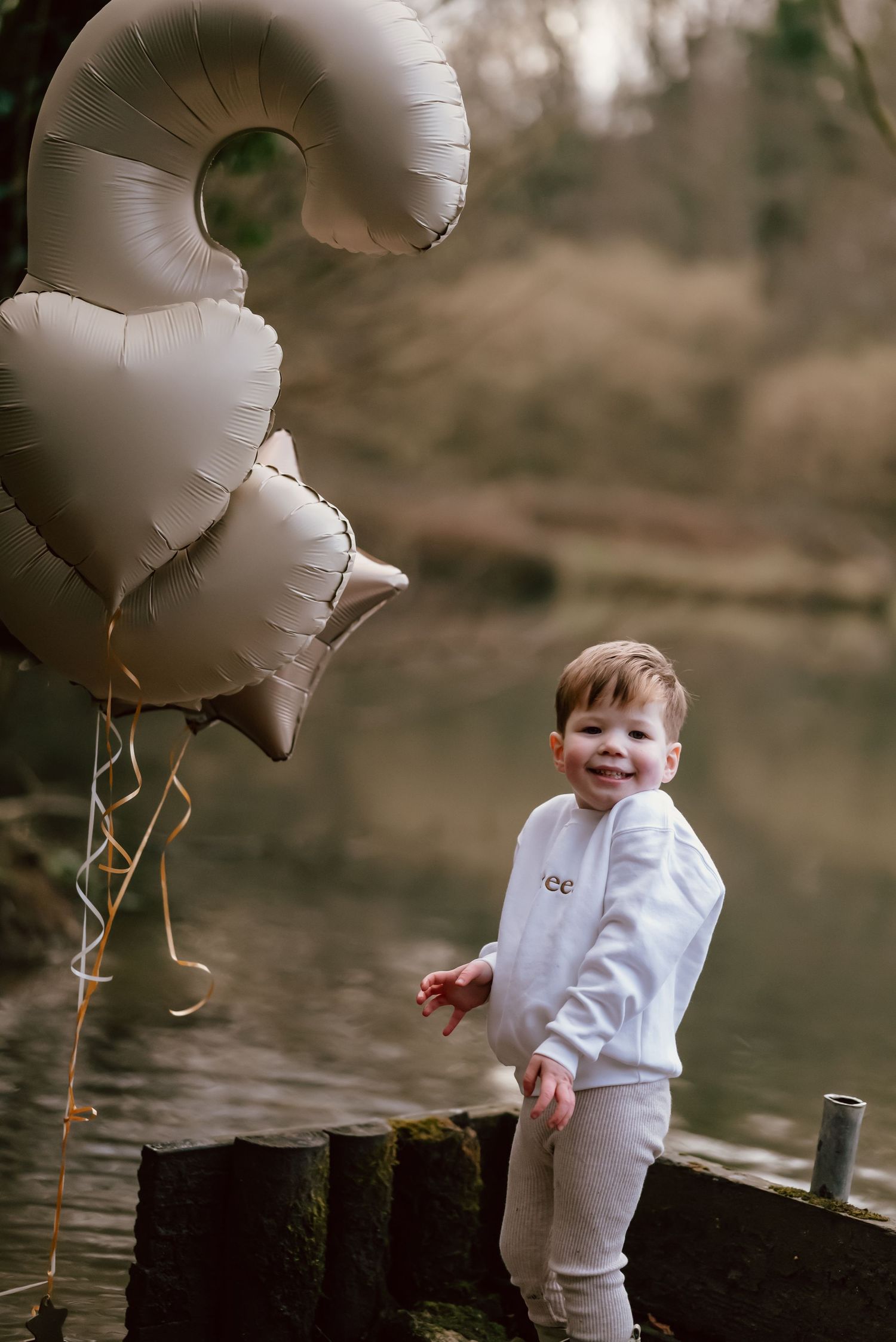 A small child stands by a lake holding white star-shaped balloons on a cloudy day.