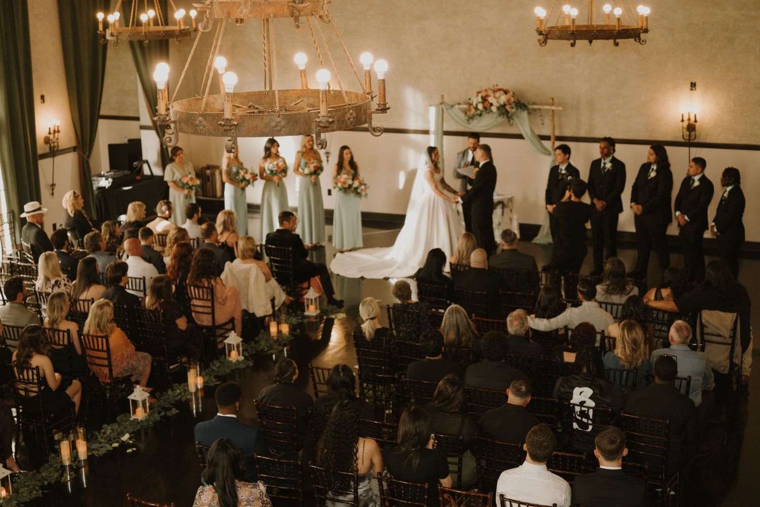 An indoor wedding ceremony with guests seated watching the couple exchange vows under chandeliers.