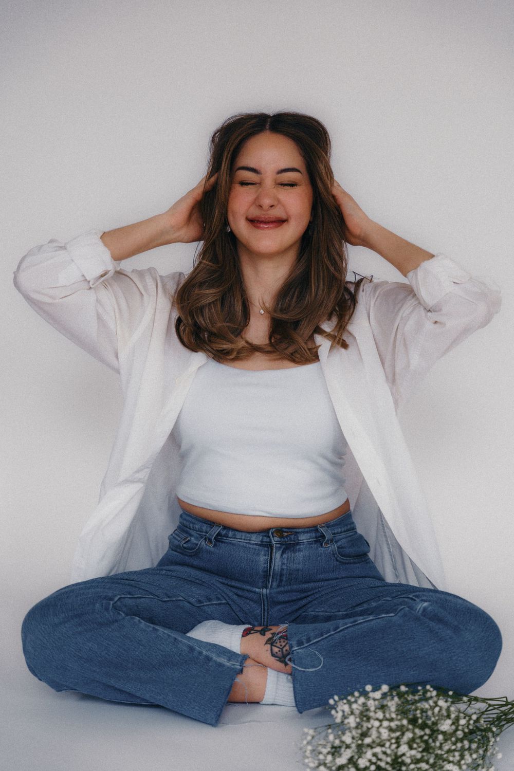 Woman in white shirt and jeans poses with baby's breath flowers against grey backdrop.