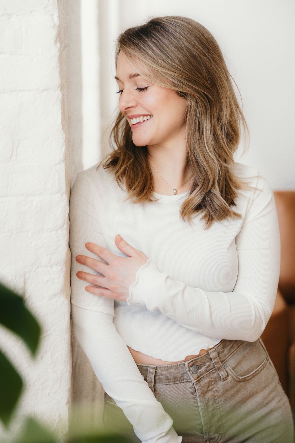 Portrait of a person in a white sweater and tan pants posing against a light wall with natural lighting.