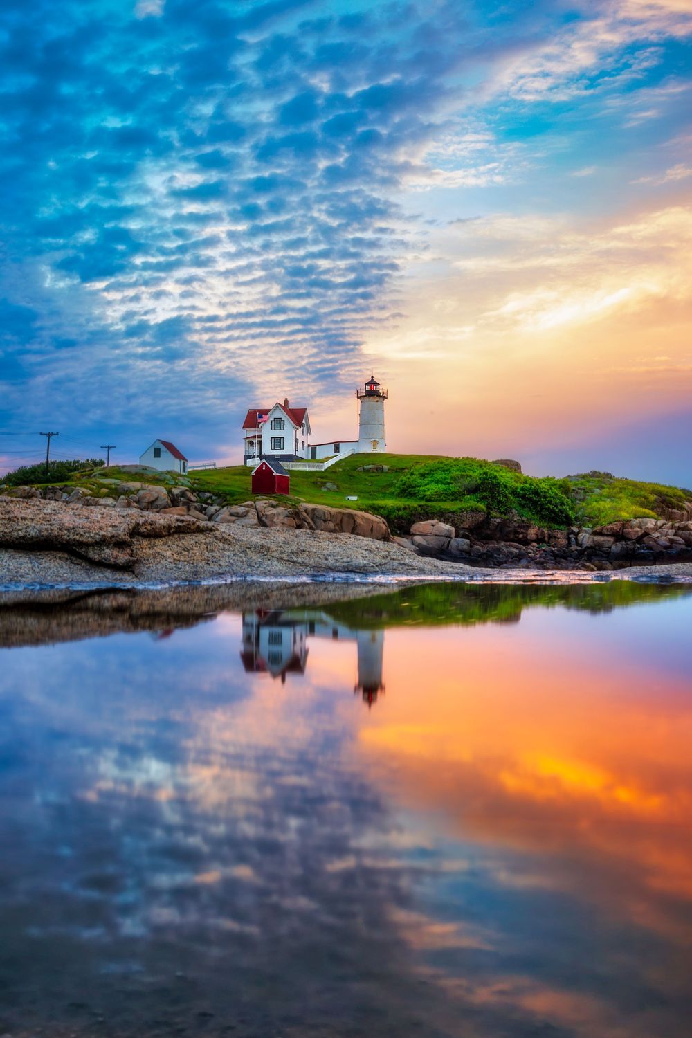 Nuble lighthouse on rocky coast reflects in tidal pool at sunset with dramatic colorful sky in York Maine.