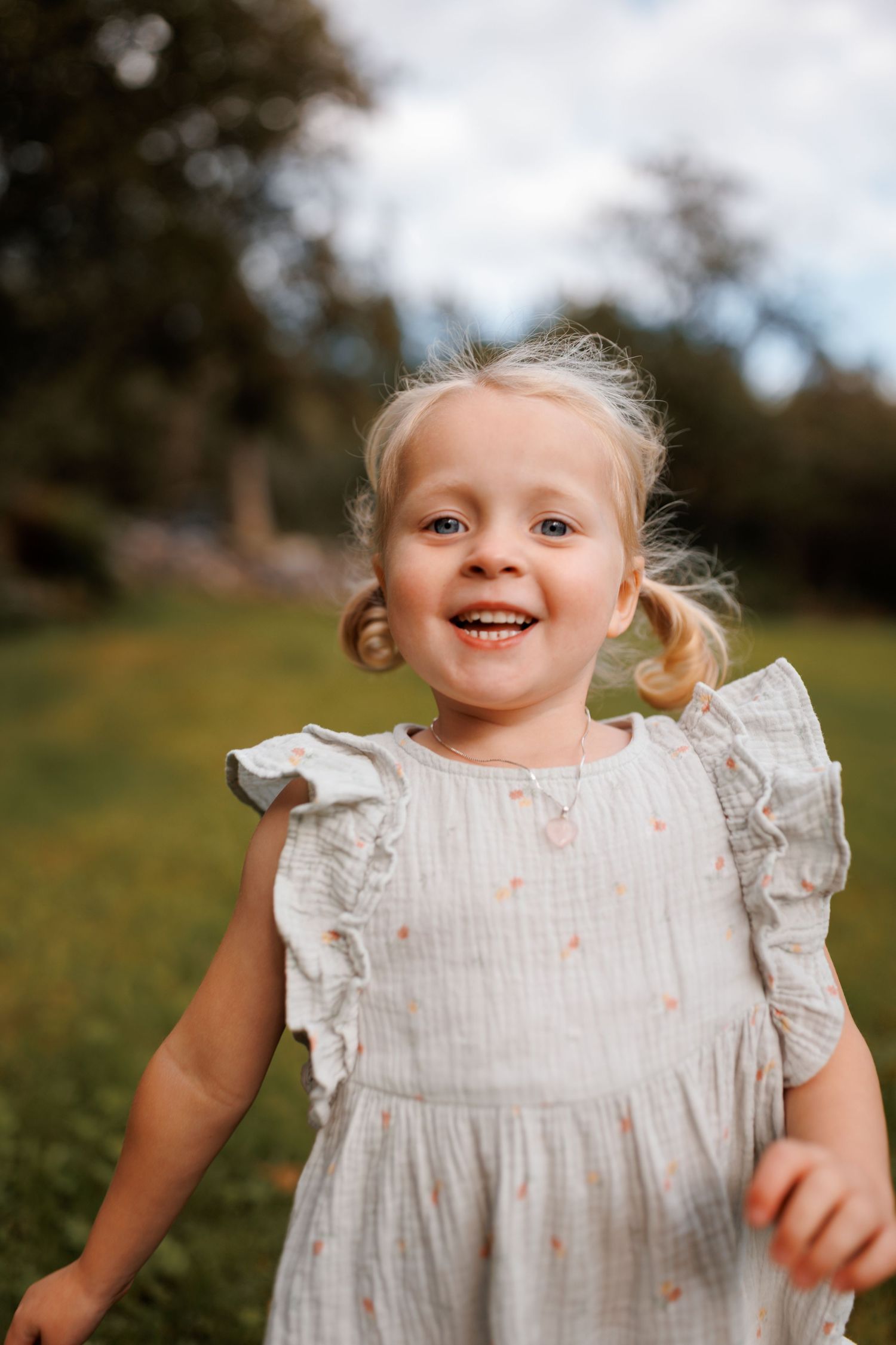 A joyful child wearing a white ruffled dress stands outdoors in a natural setting with trees in the background.