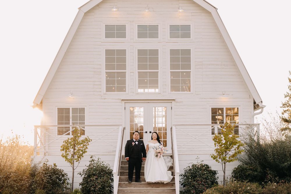 A newlywed couple poses in front of a white barn-style wedding venue at sunset.