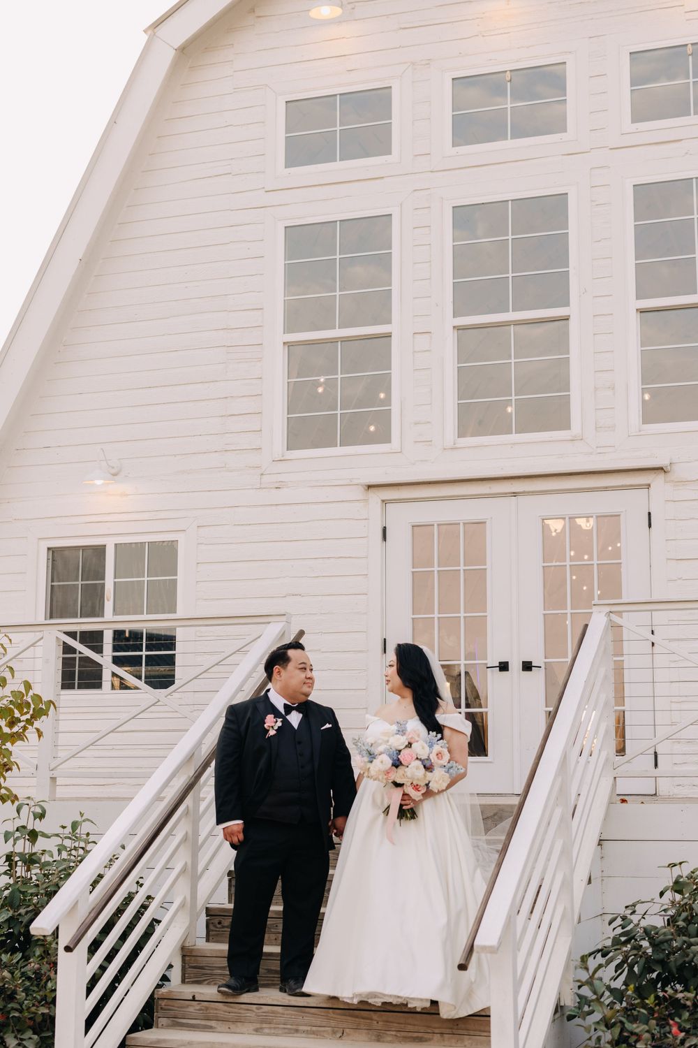 A wedding couple stands on white stairs outside a rustic barn-style venue.