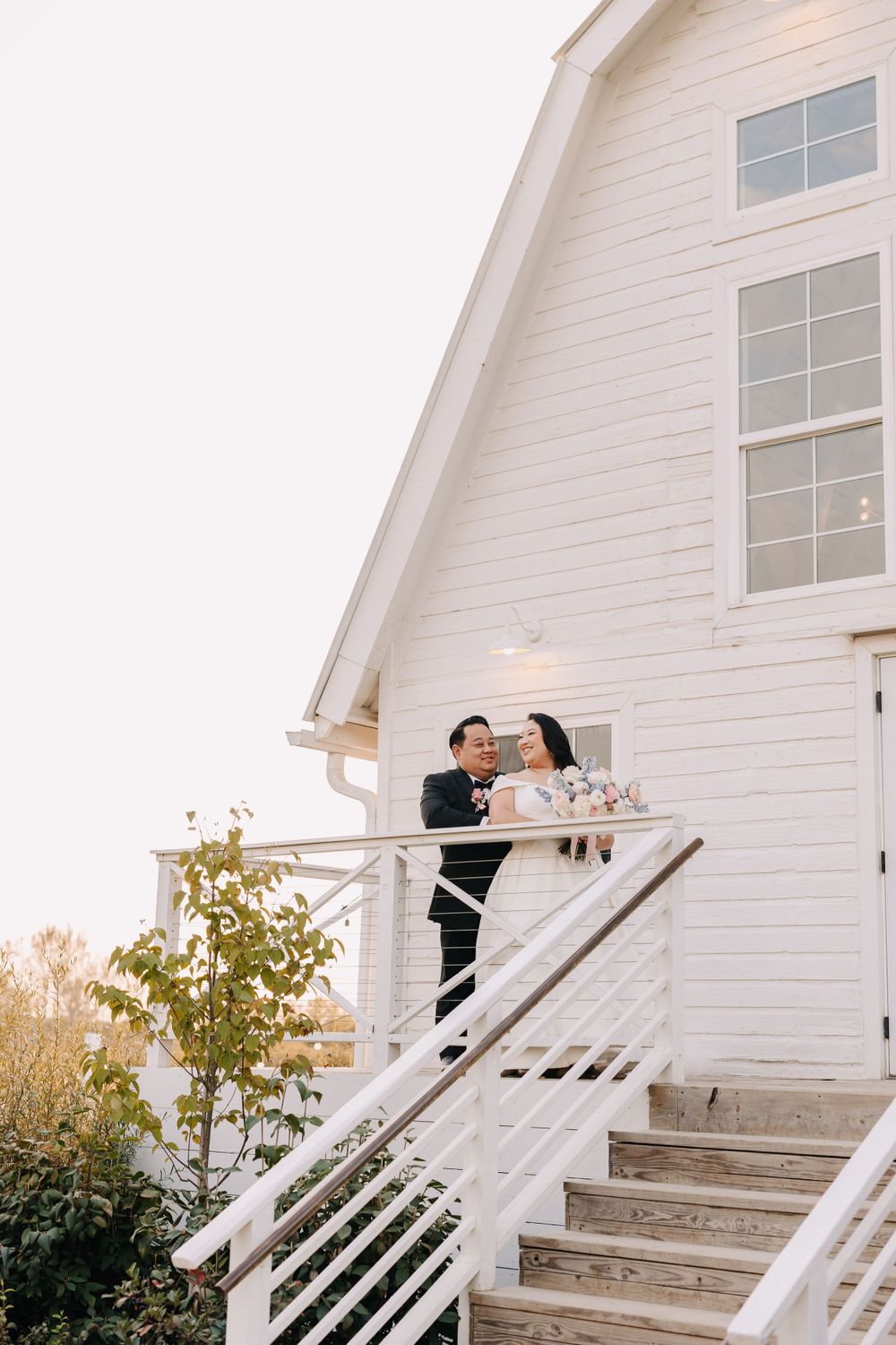 A couple embraces on the white staircase of a rustic barn venue.