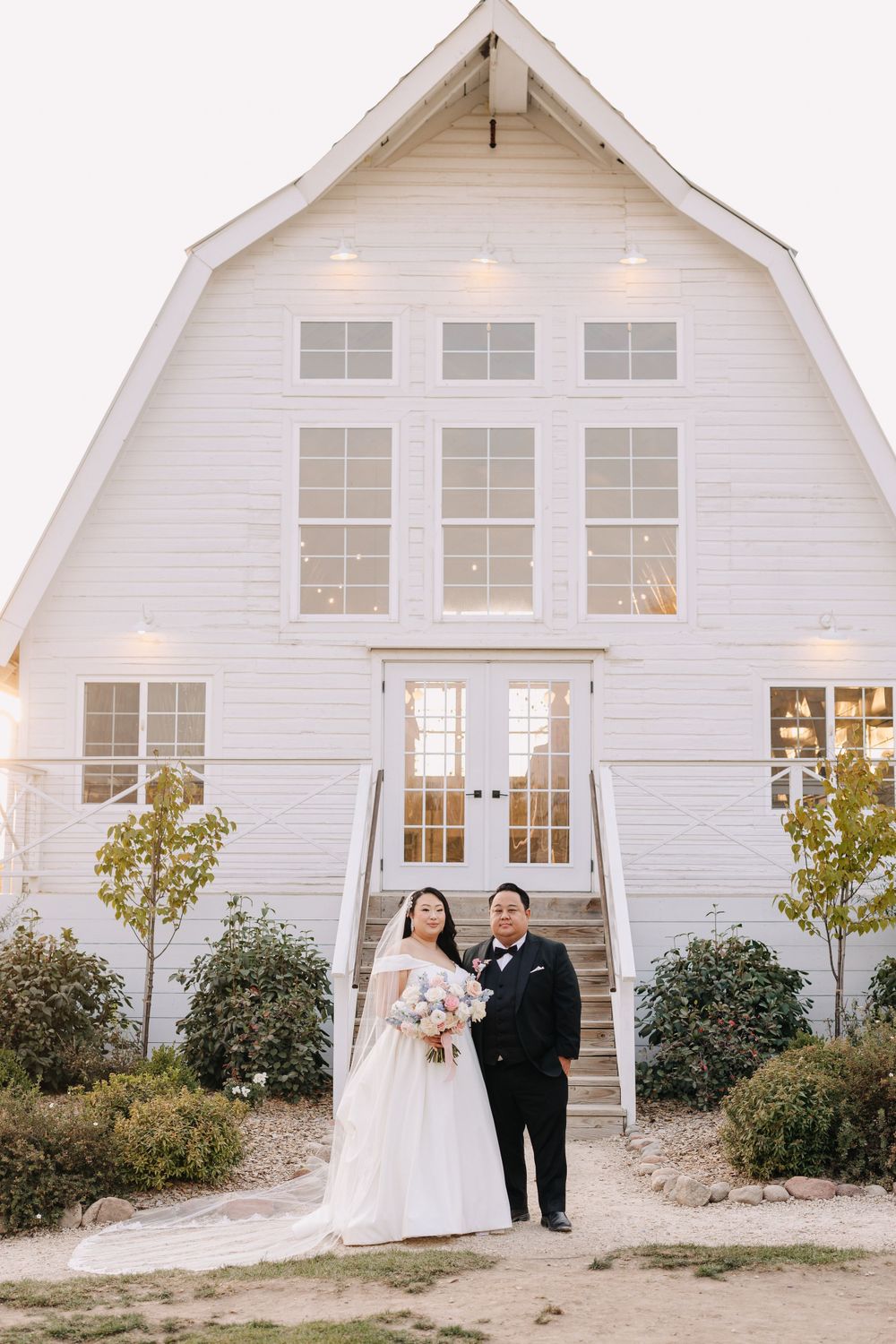 A wedding couple poses in front of a white barn-style chapel with French doors.