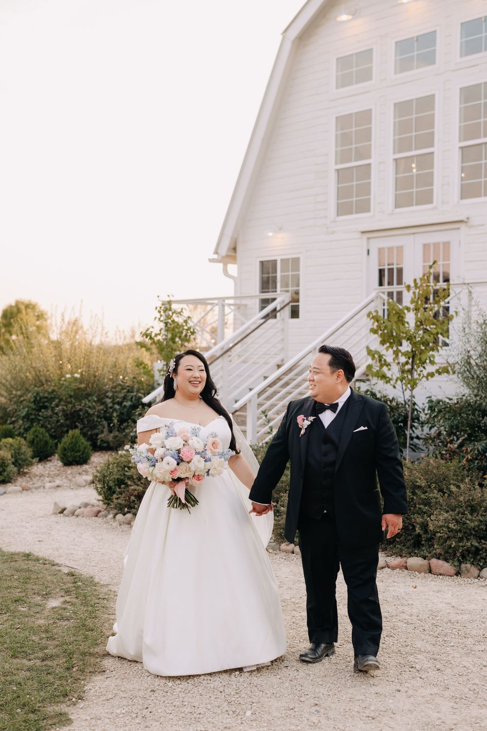 A couple walks hand in hand outside a white barn venue with pink floral bouquet.
