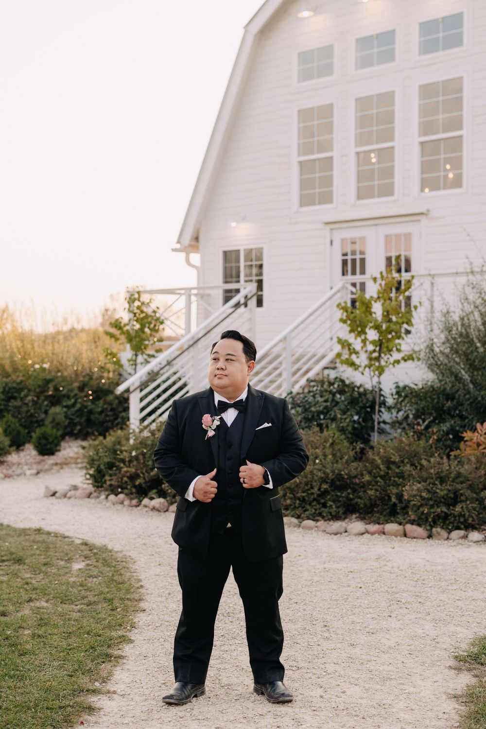 A groom in black tuxedo poses confidently in front of a white barn wedding venue.